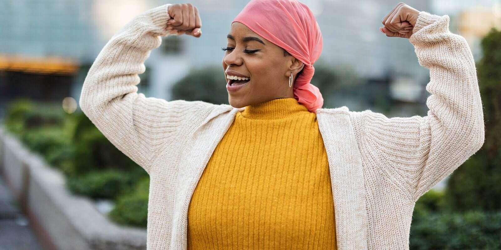 latina woman, fighting breast cancer, wears a pink scarf, and clenches her arms as a survivor fighter