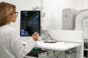 female doctor examines breast scan on monitor while patient in blue gown undergoes mammogram screening in the background