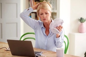 menopausal mature woman having hot flush at home cooling herself with fan connected to laptop