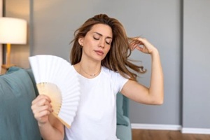 woman sitting on sofa using a white hand fan to cool down from hot flashes or high temperature at home experiencing heat wave